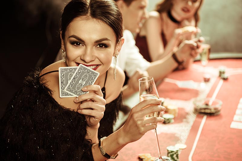 Lady sitting at poker table, holding cards in one hand and a glass of champagne in the other.
